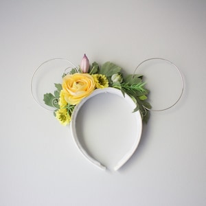 May include: White headband with a floral design featuring yellow and green flowers. The headband has two silver wire loops that resemble Mickey Mouse ears.