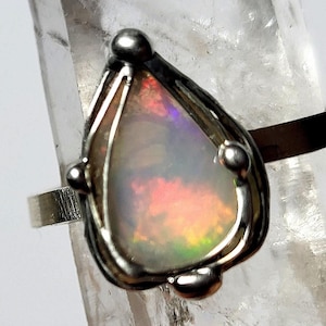 Könnte beinhalten: Ein silberner Ring mit einem tropfenförmigen Opal-Edelstein, der in einem filigranen Drahtrahmen gefasst ist. Der Opal zeigt einen Regenbogen an Farben, darunter Rosa, Orange, Gelb, Grün und Blau.