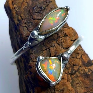 Könnte beinhalten: Ein silberner Ring mit zwei Opal-Steinen, die in einem einzigartigen, organischen Design gefasst sind. Die Opale sind in einem blassen, irisierenden Weiß mit orangefarbenen und grünen Schimmern.