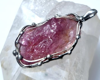 Könnte beinhalten: Ein rosa Kristall-Anhänger mit einer silbernen Drahtummantelung. Der Kristall hat eine raue, natürliche Form und glänzt sehr.
