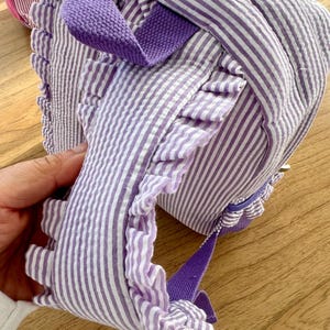 May include: Purple and white striped backpack with a ruffled trim and purple straps.