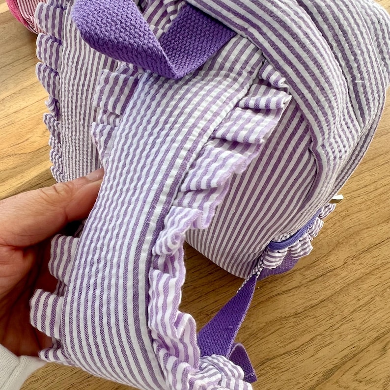 May include: Purple and white striped backpack with a ruffled trim and purple straps.