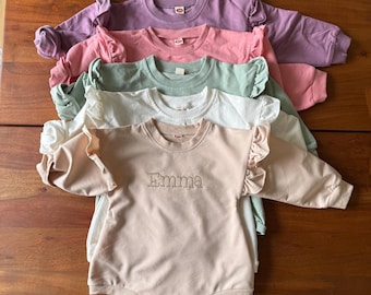 May include: A stack of five baby rompers in various pastel colors: lavender, pink, mint green, white, and beige. The beige romper has the name "Emma" embroidered on the front. Each romper features long sleeves and ruffled shoulder detailing.