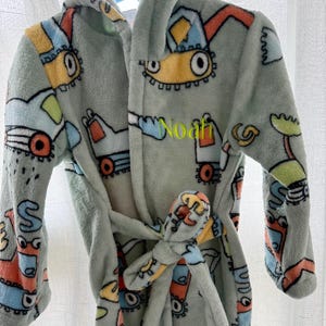 May include: A light green plush bathrobe with a hood and tie, featuring colorful cartoon construction vehicle designs. The name "Noah" is embroidered on the front. The robe is designed for children.