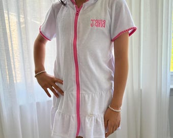 May include: White hooded terry cloth robe with pink trim and zipper. The robe has short sleeves and a ruffled hem. The name "Jayla" is embroidered in pink on the chest. The robe is designed for children.