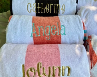 May include: White beach towels with a central stripe in black, coral, and pink. Each towel is embroidered with a name in gold or teal thread: "Catherine", "Angela", and "Jolynn". The towels are folded and stacked.