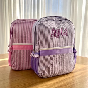 May include: Two personalized backpacks with gingham patterns. One is pink with a white gingham pattern and the other is purple with a white gingham pattern. The purple backpack has the name "Ayla" embroidered on the front.