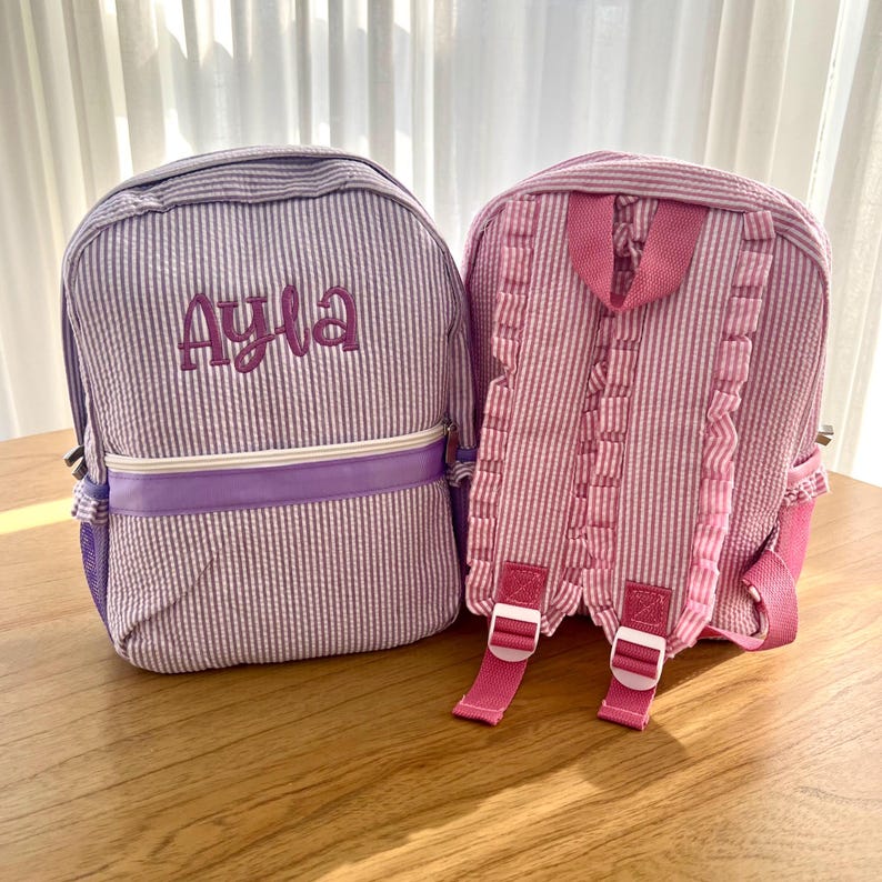 May include: Two personalized backpacks with pink and purple gingham patterns. The purple backpack has the name "Ayla" embroidered on the front.