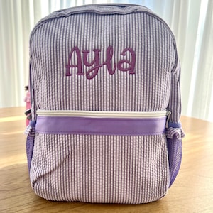 May include: Purple and white striped backpack with a purple zipper and a mesh pocket. The backpack is personalized with the name "Ayla" in purple embroidery.