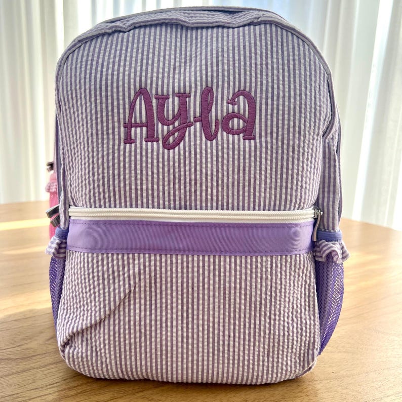 May include: Purple and white striped backpack with a purple zipper and a mesh pocket. The backpack is personalized with the name "Ayla" in purple embroidery.