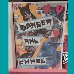 以下が含まれることがあります： 「DANGER AND CHAOS」の文字が太字で書かれた、ミクストメディアのアート作品。猫、骸骨、ピンクのハートの画像が特徴です。「13」の数字も表示されています。背景はコラージュのような外観です。