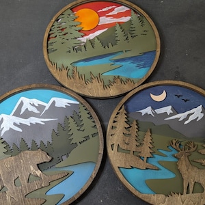 Peut inclure: Trois décorations murales circulaires en bois superposé. La pièce du haut montre un coucher de soleil sur un lac et une forêt. La pièce en bas à gauche représente un ours dans une forêt avec des montagnes. Celle de droite montre un cerf.