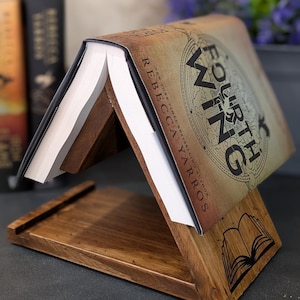 May include: A wooden book stand holding a book titled "Fourth Wing" by Rebecca Yarros. The stand is angled, with a tray at the base and a book icon etched on the side. The book's cover is visible, displaying a map design.
