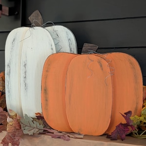 May include: Two wooden pumpkin decorations, one white and one orange, with wire stems and brown accents. The pumpkins are placed on a wooden surface with autumn leaves, ideal for seasonal decor.