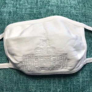 May include: White face mask with a white embroidered design of a building with a spire on the front.