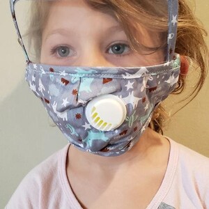 以下が含まれることがあります： 透明なプラスチックシールドと白いバルブが付いた灰色のフェイスマスクを着用した子供。マスクには星と漫画の動物の模様があります。子供は虹のデザインのピンクとグレーのシャツを着ています。