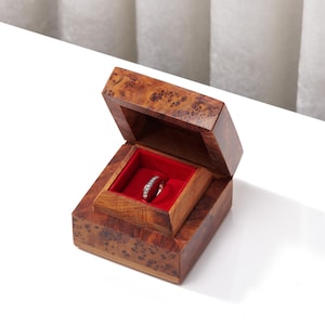 May include: A polished wooden ring box with a distinctive burl grain pattern and a hinged lid. The interior features a vibrant red velvet lining that holds a silver-toned band embedded with a series of small, clear, circular stones along its top.