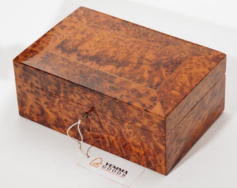 May include: A wooden box with a burl wood finish. The box has a brass lock and keyhole. The box is closed and has a tag attached to it that says "Yemma Goods".