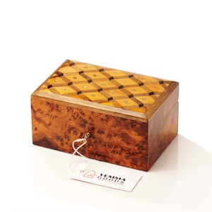 May include: A wooden box with a geometric inlay design on the lid. The box is made of a dark wood with a light brown finish. The box has a keyhole and a small tag attached to it that says "Yemma Goods The Moroccan Home".