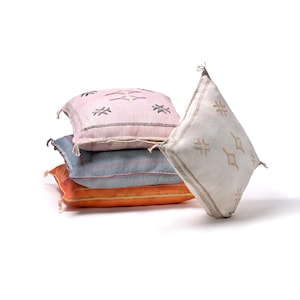 May include: A stack of square decorative pillows in various colors. The pillows include a light pink, blue, and orange pillow. The light pink pillow has black embroidered designs. The white pillow has brown embroidered designs.