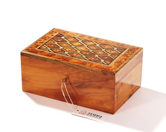May include: A wooden box with a decorative inlaid pattern on the lid. The box is made of light brown wood and has a brass latch. The box is sitting on a white surface. The box has a tag attached to it that says "Yemima Goods".