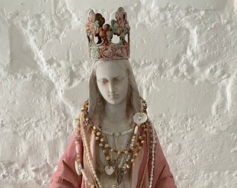 May include: A white statue of a woman wearing a pink robe and a crown. She is adorned with multiple necklaces, including a beaded necklace, a shell necklace, and a chain necklace with a heart pendant.