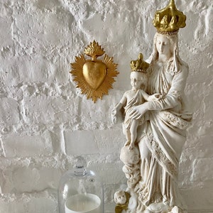May include: White ceramic statue of the Virgin Mary holding a baby Jesus. The statue is adorned with gold accents and a crown. The statue is displayed on a white shelf with a gold heart ornament and glass orbs.