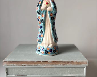 May include: A ceramic figurine of a woman in a blue and floral patterned robe, holding a heart. The figurine is on a light blue, distressed wooden stand with four rounded legs. The figurine has a white dress and a red heart.