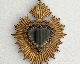 May include: A gold-coloured, heart-shaped wall mirror with a sunburst design. The mirror is framed with small, clear stones and has a loop for hanging. The mirror is suspended from a thin, brown ribbon.