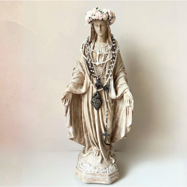 Puede incluir: Estatua crema de una figura religiosa, adornada con una corona floral y m&uacute;ltiples hilos de collares de cuentas. La estatua tiene una expresi&oacute;n serena y est&aacute; sobre una base decorativa. Un colgante en forma de coraz&oacute;n cuelga de los collares.