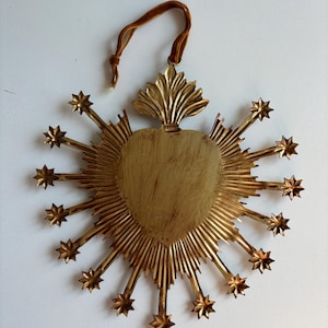 May include: A gold-colored, heart-shaped wall hanging with radiating lines and star accents. It features a velvet ribbon for hanging. The heart has a textured surface and is surrounded by starburst rays.