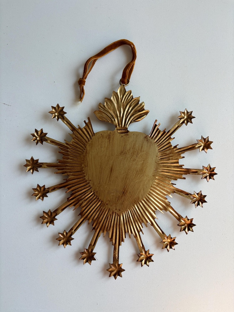 May include: A gold-colored, heart-shaped wall hanging with radiating lines and star accents. It features a velvet ribbon for hanging. The heart has a textured surface and is surrounded by starburst rays.