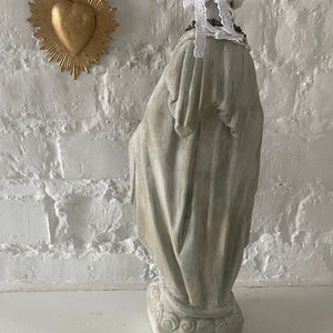 May include: A white painted plaster statue of a woman wearing a white lace crown and a beaded necklace. The statue is standing on a base with a decorative design. The statue is in front of a white brick wall.