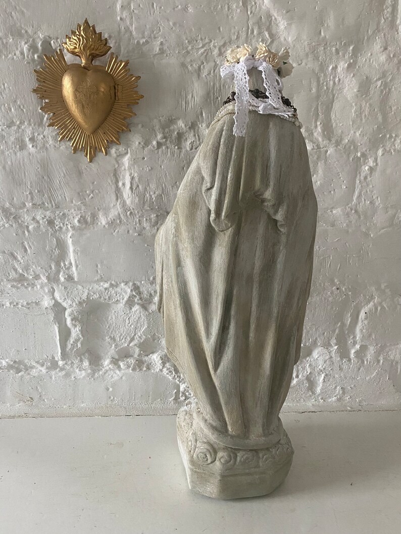 May include: A white painted plaster statue of a woman wearing a white lace crown and a beaded necklace. The statue is standing on a base with a decorative design. The statue is in front of a white brick wall.