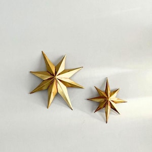 May include: Two gold-colored star-shaped decorative objects. The larger star has eight points, and the smaller star has seven points. Both stars are made of a shiny, metallic material and are set against a white background.