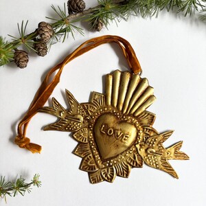 May include: A gold-colored metal heart-shaped ornament with the word "LOVE" embossed on it. The ornament features a sunburst design and two bird figures. A gold ribbon is attached for hanging. Decorative pine branches and pinecones are in the background.
