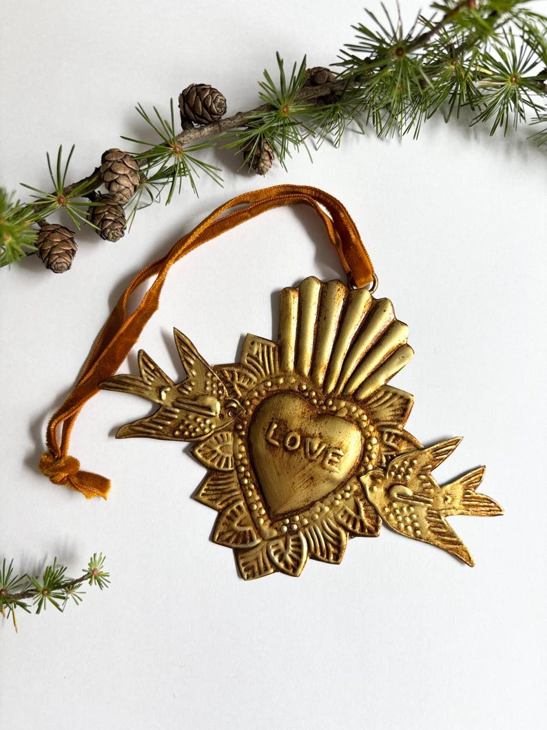 May include: A gold-colored metal heart-shaped ornament with the word "LOVE" embossed on it. The ornament features a sunburst design and two bird figures. A gold ribbon is attached for hanging. Decorative pine branches and pinecones are in the background.
