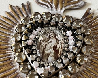 May include: A decorative wall hanging in the shape of a sacred heart, featuring a central oval image of a figure with a heart, surrounded by small white floral accents, clear rhinestones, and a gold-toned metal sunburst frame with a top loop.