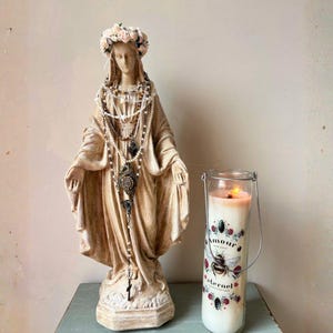 Puede incluir: Una estatua beige de la Virgen Mar&iacute;a adornada con una corona floral y collares en capas. Una vela encendida en un frasco de vidrio con una etiqueta decorativa con una abeja y rosas est&aacute; al lado de la estatua.
