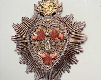 May include: A heart-shaped wall decoration with a metallic finish. It features faux jewels, coral-coloured roses, and a small religious image. The design includes a gold flame and sunburst rays, creating a unique, ornate piece. The measurements are in centimetres.