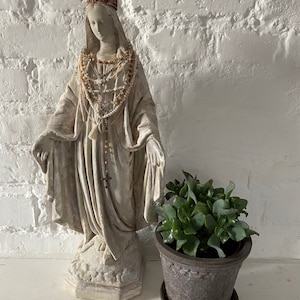 Puede incluir: Una estatua de yeso blanco de la Virgen María con una corona dorada y un collar de cuentas. La estatua está de pie sobre una base blanca y está frente a una pared de ladrillo blanco. Una pequeña planta en maceta está en primer plano.