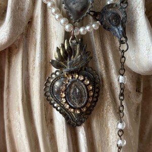 Puede incluir: Un collar de estilo vintage con un colgante en forma de coraz&oacute;n con una imagen religiosa central, rodeado de detalles brillantes. El colgante est&aacute; unido a un collar de perlas y una cadena con peque&ntilde;as cuentas transparentes. Aspecto envejecido.