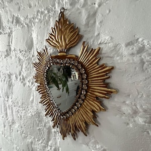 Könnte beinhalten: Ein goldener herzförmiger Spiegel mit einem Flammenmuster oben und Strasssteinen am Rand. Der Spiegel hängt an einer weißen Wand.