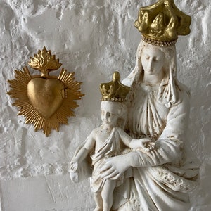 May include: White plaster statue of the Virgin Mary and baby Jesus. Mary is wearing a gold crown with rhinestones and a white robe. Jesus is wearing a gold crown with rhinestones and a white robe. The statue is on a white background. There is a gold heart with a cross on it in the background.