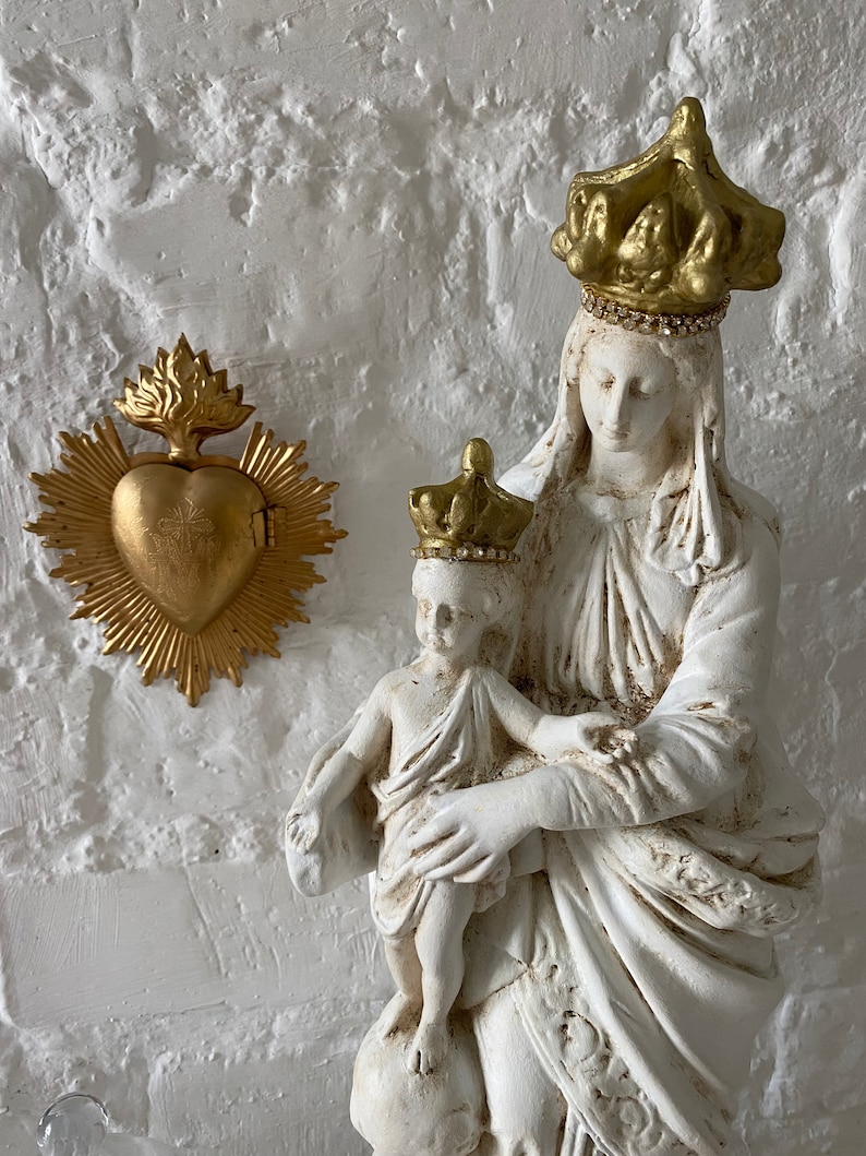May include: White plaster statue of the Virgin Mary and baby Jesus. Mary is wearing a gold crown with rhinestones and a white robe. Jesus is wearing a gold crown with rhinestones and a white robe. The statue is on a white background. There is a gold heart with a cross on it in the background.
