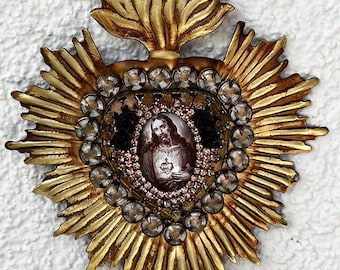 May include: A gold-toned metal sacred heart ornament featuring a central oval portrait of a figure with a heart, surrounded by a border of small clear rhinestones and larger faceted crystals, topped with a stylised flame motif and a hanging loop.
