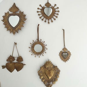 May include: A collection of antique gold-colored wall mirrors in various shapes, including hearts and sunburst designs. Some mirrors have a heart-shaped frame, while others feature a round or oval mirror. The mirrors are hung on a white wall.