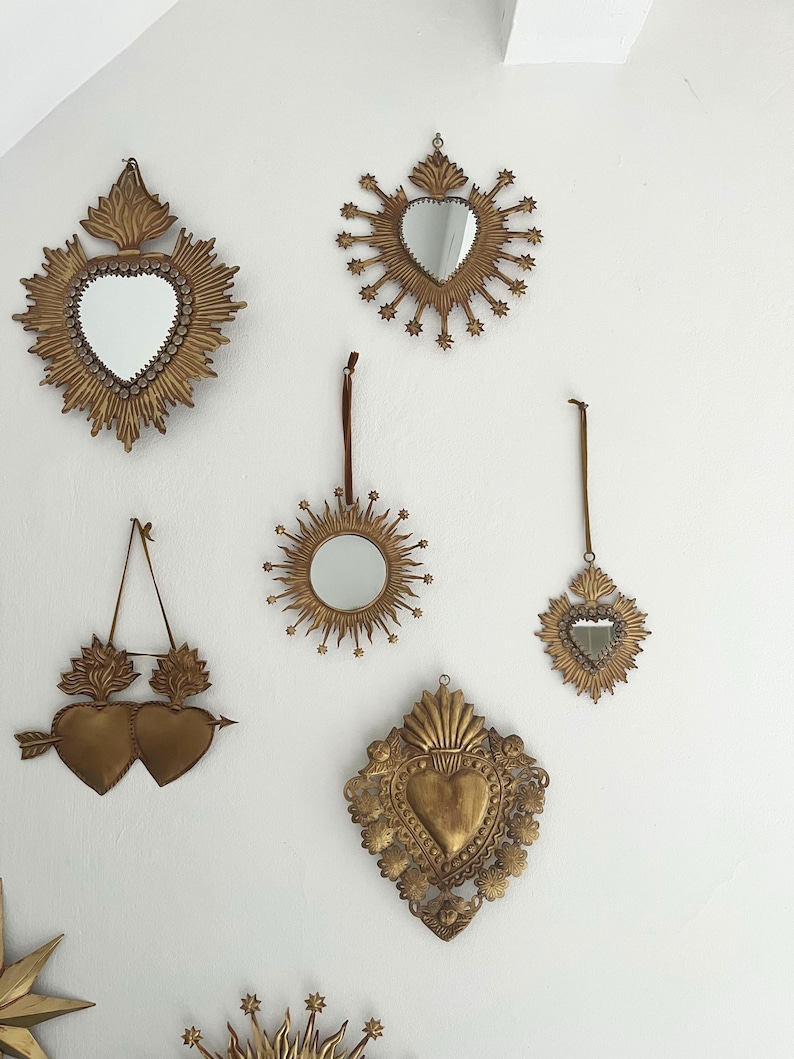 May include: A collection of antique gold-colored wall mirrors in various shapes, including hearts and sunburst designs. Some mirrors have a heart-shaped frame, while others feature a round or oval mirror. The mirrors are hung on a white wall.