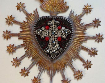 May include: An elaborate, heart-shaped religious pendant featuring a central cross. The heart is black with red detailing and a sparkling cross. Gold-toned rays and starbursts radiate from the heart, with a brown cord for hanging.