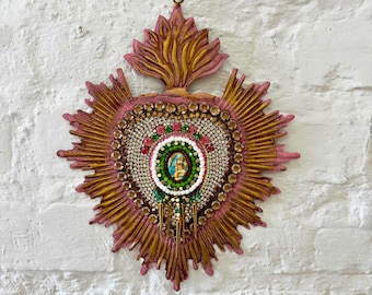May include: A heart-shaped wall hanging with a central religious image. The design features a sunburst pattern in pink and gold, adorned with rhinestones and beads. A distinctive piece of religious art, suitable for home decor. The piece is mounted on a white brick wall.
